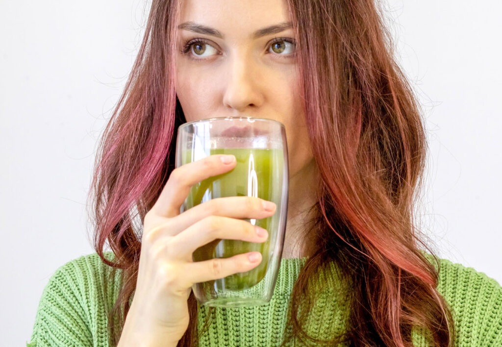 woman with brown and pink hair drinking matcha from a glass wearing a green sweater