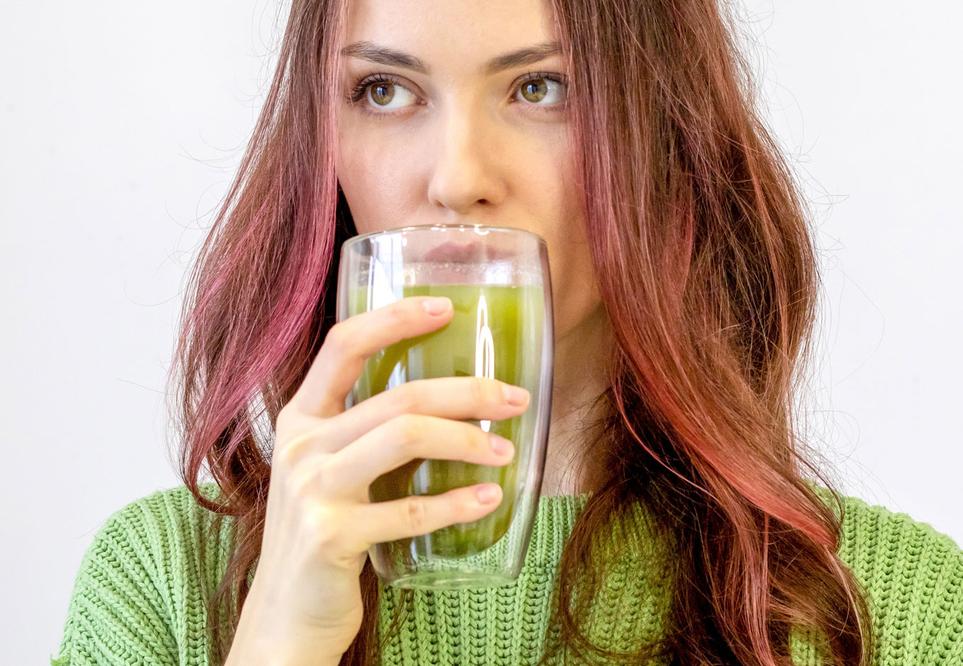 woman with brown and pink hair drinking matcha from a glass wearing a green sweater