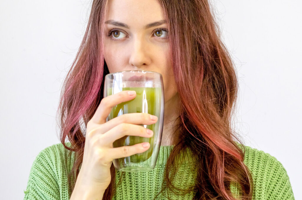 woman with brown and pink hair drinking matcha from a glass wearing a green sweater
